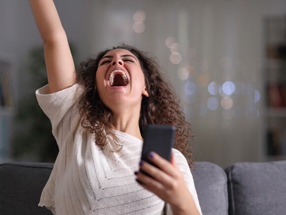 אישה צעירה מחזיקה טלפון בידה ומרימה את ידה מעלה בשמחה
