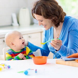 אמא ותינוק יושבים ליד שולחן בעוד האמא מחייכת לתינוקה ומחזיקה בידה כפית מלאה באוכל לארוחת בוקר