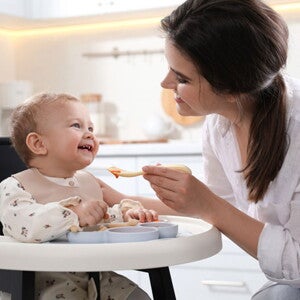 אמא מאכילה את תינוקה בכפית, כשהוא יושב בכסא האוכל שלו