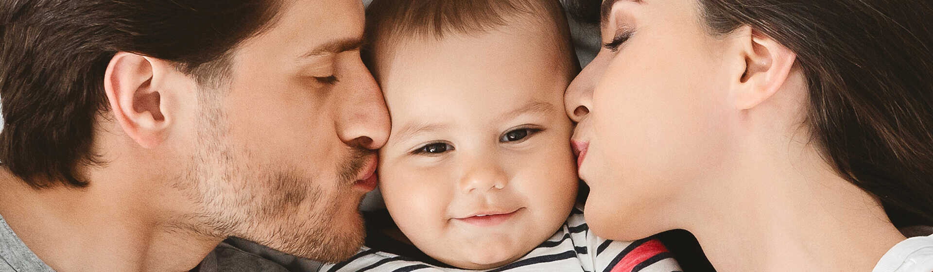 זוג הורים מנשקים עם תינוקם משני צידיו כשהוא שמח במרכז